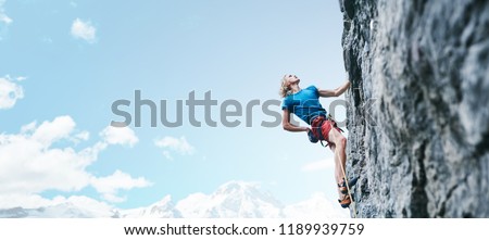 man rock climber with long hair. side view of young man rock climber in bright red shorts climbing the challenging route on the cliff on the blue sky background. rock climber climbs on a rocky wall.