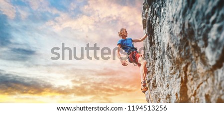 man rock climber with long hair. side view of young man rock climber in bright red shorts resting while climbing the challenging route on the cliff on the colorful down sky background. rock climber