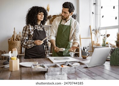 Man rolls clay with rolling pin while woman stands next to him holding stack in hands. Pottery studio with various tools and materials around them, creating handmade ceramic art together. - Powered by Shutterstock