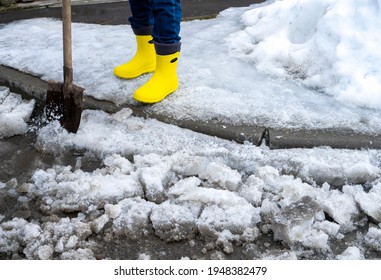 Man In Rubber Foam Boots Cleaning The Track From Old Snow. He Shovels Off Dirty Wet Snow With A Shovel So That It Melts Faster