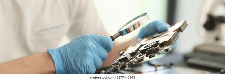 Man in rubber gloves looks at printed circuit board of laptop through magnifier glass. Electronic devices repair service and maintenance - Powered by Shutterstock