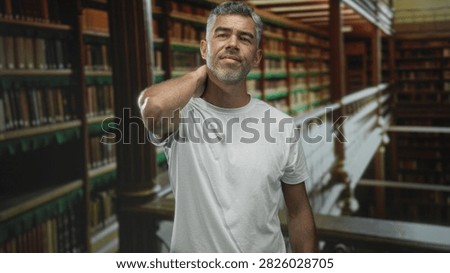 Man rubbing neck in a library between tall bookshelves, wearing a white tshirt and wincing with hand pressed to back of neck; discomfort.