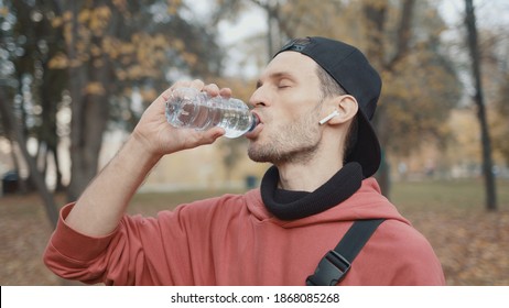 Man Runner Closeup In Red Hoodie And Earbuds Drinking Water In The Park Slow Motion, On Background Of Yellow Trees. Gimbal Shot Of Man Drinking Water From The Bottle