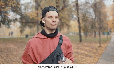 Man Runner Closeup In Red Hoodie And Earbuds Drinking Water In The Park Slow Motion, On Background Of Yellow Trees. Gimbal Shot Of Man Drinking Water From The Bottle