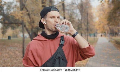 Man Runner Closeup In Red Hoodie And Earbuds Drinking Water In The Park Slow Motion, On Background Of Yellow Trees. Gimbal Shot Of Man Drinking Water From The Bottle