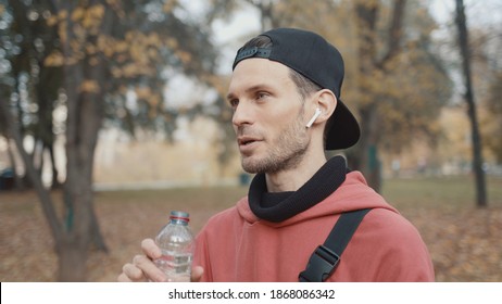 Man Runner Closeup In Red Hoodie And Earbuds Drinking Water In The Park Slow Motion, On Background Of Yellow Trees. Gimbal Shot Of Man Drinking Water From The Bottle