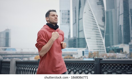 Man Runner In Red Hoodie On Background Of Business Skyscrapers In Moscow, Slow Motion. Middle Shot Of Young Man Listening To Music Jogging In The City Business Area