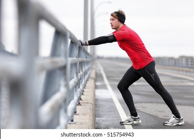 Man Runner Stretching Legs Before Exercise Run Outdoors In City Urban Street During Winter. Athlete Wearing Smartwatch, Phone Armband For Music App And Warm Gloves, Headband, Long Tights Underwear.