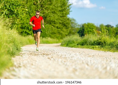 Man runner trail running on country road in summer sunset. Young athlete male cross country training and doing workout outdoors in green nature, summer blue sky
