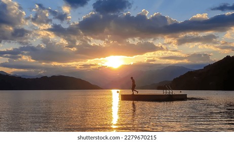A man running along wooden platform drifting on the MIllstaetter lake during the sunset. Jumping into deep water. The sun sets behind high Alps. Calm surface of the lake reflects the orange sky. Fun - Shutterstock ID 2279670215