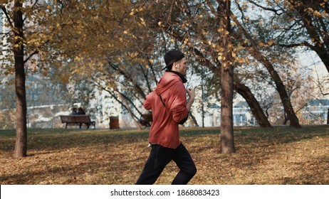 Man Running In The Park Slow Motion, Young Man In Sport Clothes And Cap Listening To Music Running Through The Park In The Autumn Day, Middle Shot