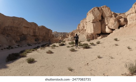 Man running through a desert landscape with rocky formations and sparse vegetation under a clear blue sky, capturing an adventurous spirit in a barren wilderness - Powered by Shutterstock