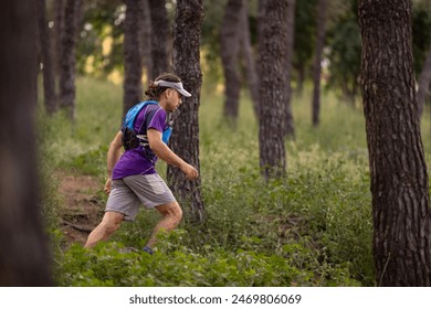 A man is running through a forest with a backpack on. The man is wearing a purple shirt and gray shorts