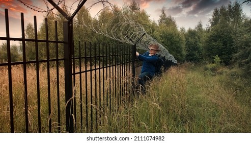 Man Runs To The Fence With Barbed Wire And Climbs On It. Evening Sunset. The Concept Of Illegal Border Crossing, Penetration Into A Protected Object And Unlawful Migration