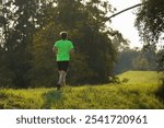 A man runs on a grassy path in a lush green park, wearing a neon green shirt and black shorts. A calm natural setting, the beauty and tranquility of exercising outdoors