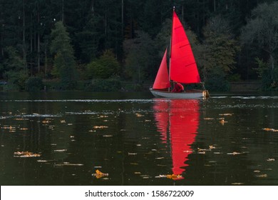Man Sailing Dinghy With Red Sail On Autumn Lake