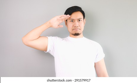 A Man In Salute Pose And Serious Face. An Asian Man With White T-shirt And Grey Background.