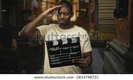 Man salutes with right hand while holding a clapperboard in a narrow urban street with shuttered storefronts and stone steps; confidence cinema.