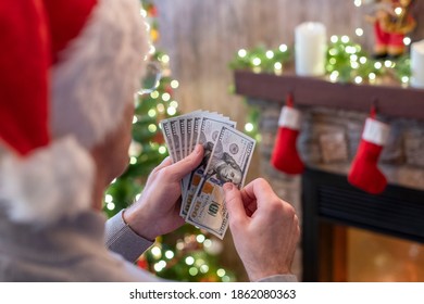 Man in a Santa claus hat counting american dollars sitting near christmas tree and fireplace. Spending money on gift at christmas time. concept. - Powered by Shutterstock