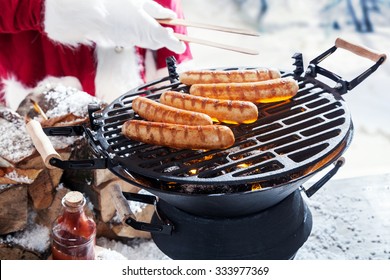 Man in Santa outfit grilling sausages outdoors in the snow on a portable barbecue, close up of his hand in a Christmas party concept