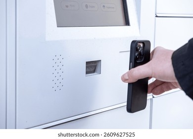Man scanning smartphone at white parcel locker screen to collect package using QR code or app during modern contactless pickup process