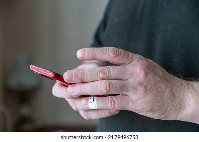 A Man Is Scrolling Through Messages On His Smartphone. A Red Smartphone Is Held In His Hands. The First Phalanx Of His Pinky Finger Is Wrapped In White Ribbon. A Smiling Face Is Painted On The Bandage