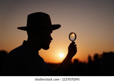 Man Searching Using Loupe With Sun On Background And Urban View. Male Detective Or Investigator Silhouette Portrait In Hat Looking With Magnifying At Side. Investigation Or Inspection Concept