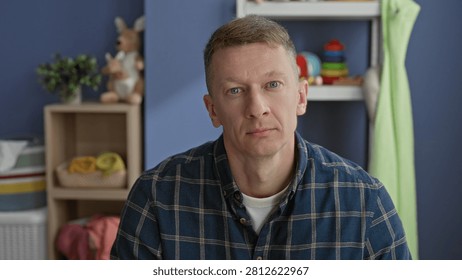 Man seated facing camera with visible face and neutral gaze, wearing plaid shirt amid toys and shelf in a nursery inside building; contemplation.