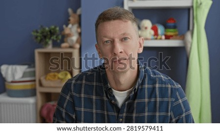 Man seated facing camera with visible face and neutral gaze, wearing plaid shirt amid toys and shelf in a nursery inside building; contemplation.