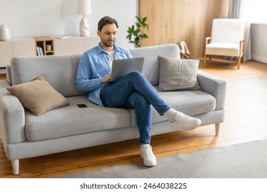 A man is seated on a couch, focused on his laptop screen. He appears engaged in his work or activities online, with a serious expression. The room is simple, with no other visible people - Powered by Shutterstock