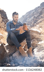 Man seated on a rock ans texting as he looks at his cell phone, he is sitting in the mountains