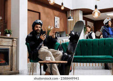 A man seated on sofa browses on his digital phone with snowboard nearby. Caucasian visitor dressed in snow clothing surfing the net using his smartphone. Winter vacation anticipation captured. - Powered by Shutterstock