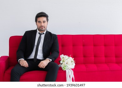 Man Seeking For Couple Concept, Handsome Caucasian Man With Beard Take Photo With Red Sofa Bed Look To Window, Groom In Tuxedo And Flower Bouquet Wafting For Perfect Girl Or Bride