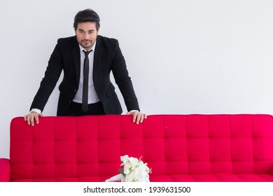 Man Seeking For Couple Concept, Handsome Caucasian Man With Beard Take Photo With Red Sofa Bed Look To Window, Groom In Tuxedo And Flower Bouquet Wafting For Perfect Girl Or Bride