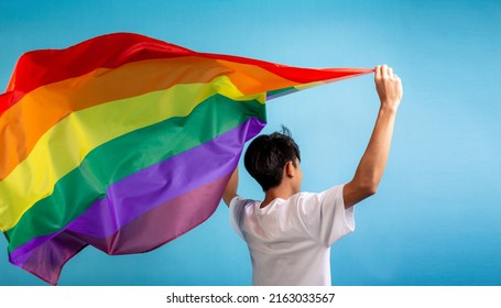 A man seen from behind holding colorful rainbow flag on blue color background. Male with a gay pride flag in studio, LGBT concept. - Powered by Shutterstock