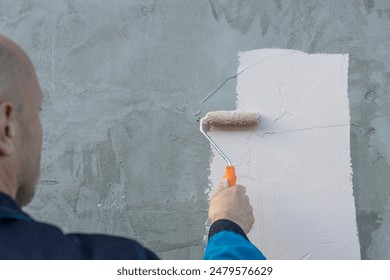 A man is seen from behind, using a paint roller to apply a coat of fresh white paint to a concrete wall, a clean, smooth stroke defining the new paint.