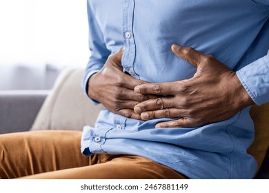 A man is seen clutching his stomach, indicating abdominal pain or discomfort. He is sitting down and appears to be in distress, suggesting a possible gastrointestinal issue or stomach ache. - Powered by Shutterstock