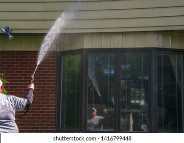 Man Is Seen With A Spray Nozzle In His Hand. He Is Spraying Water To Clean The Siding And Bricks On The Front Of A House. The House Has A Large Bay Window