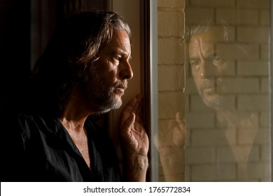 Man In Self Isolation, Stay Home Concept, Thinking Man's Reflection In Glass Window Of Brick House, Concerned Long Haired Male With Beard Looking Through Window