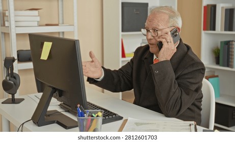 Man senior grey haired holding smartphone to ear and typing on desktop computer keyboard at office desk in a building with shelves and paperwork; focused professionalism.