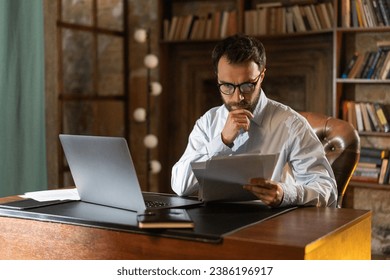 A man with serious look holds and reads important documents attentively, sits at a table in the library and uses a laptop. A respectable businessman acquainted with a lucrative contract. A young