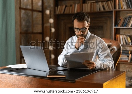 A man with serious look holds and reads important documents attentively, sits at a table in the library and uses a laptop. A respectable businessman acquainted with a lucrative contract. A young