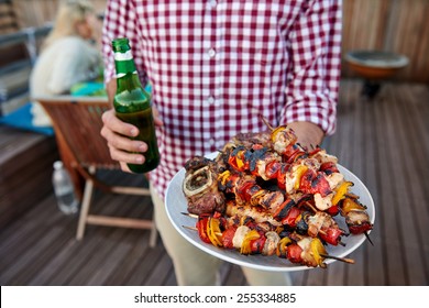 Man Serves Skewer Kebabs For Outdoor Barbeque Dinner Party Holding Beer