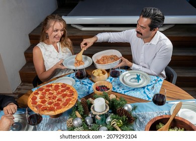 Man Serving Food To His Partner During Christmas Dinner, Sitting Joyfully At A Table With Christmas Decoration