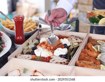 Man Serving Himself From A Boxed Platter Of Greek Food At An Outdoor Party