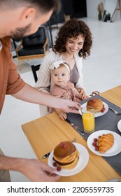 Man Serving Pancakes To Happy Wife With Infant Daughter During Breakfast