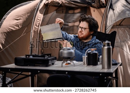Man sets up a folding windshield on a camping table with cooking equipment, including a kettle and gas canister, outside a tent.