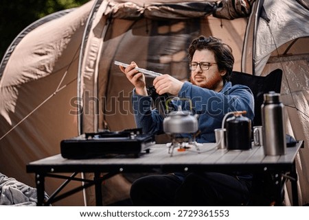 Man sets up a folding windshield on a camping table with cooking equipment, including a kettle and gas canister, outside a tent.