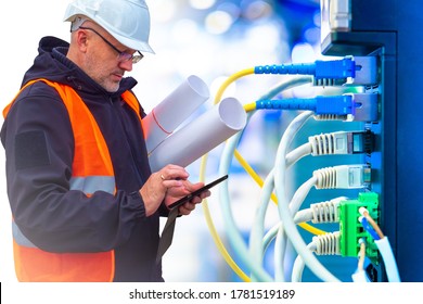 Man sets up a local network. Worker next to network equipment. System administrator in a helmet. Man holds blueprints and looks for something on phone. Server hardware setup. Wires go to the server