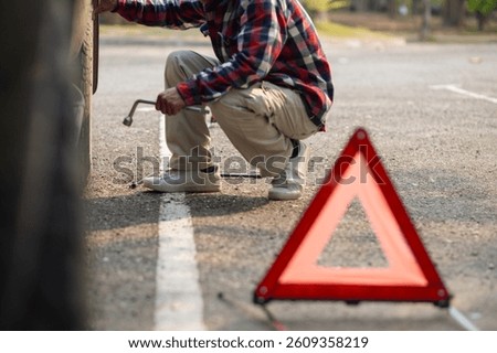 A man sets up a reflective triangle to warn other drivers to be careful of cars parked on side of the road. A man sets up an orange reflective triangle on the side of the road to change a flat tire.